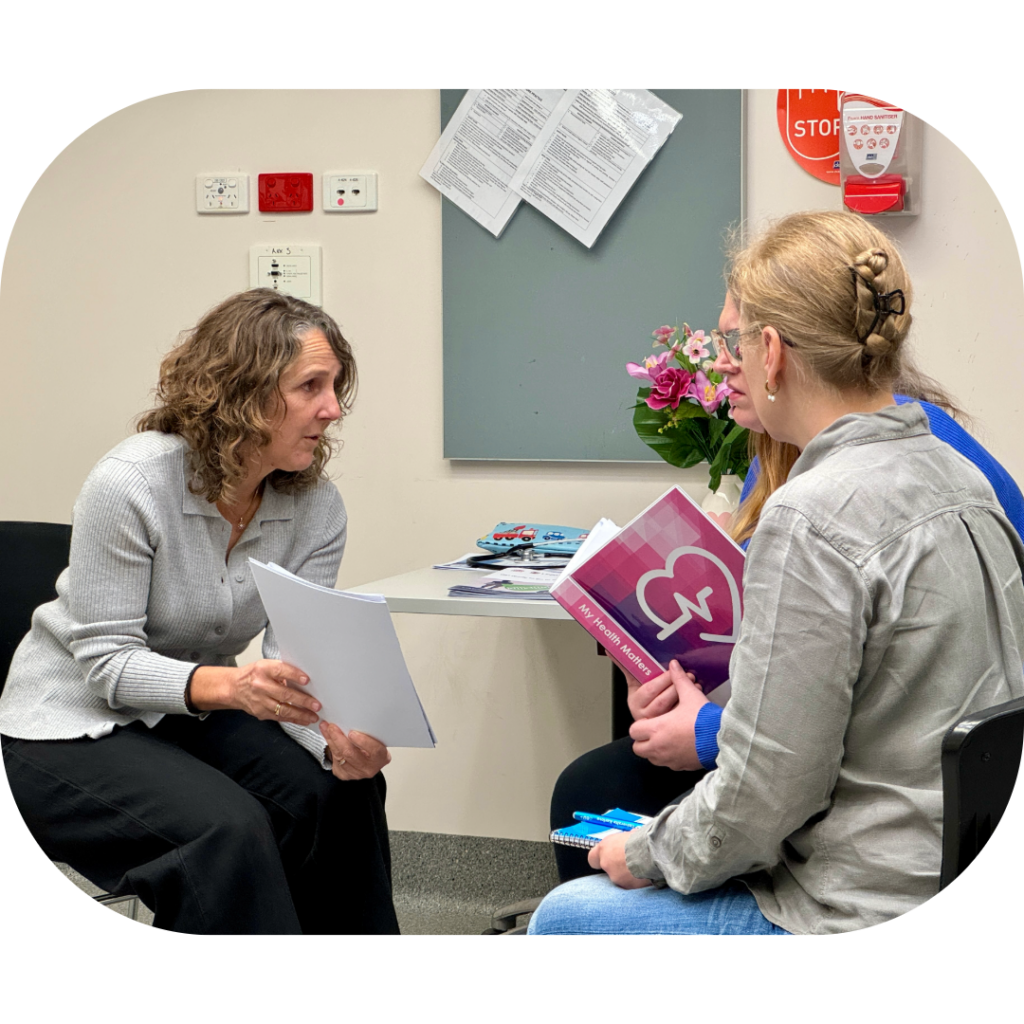 A genetic counsellor holding a booklet and speaking with her client, while a support person sits beside the client.